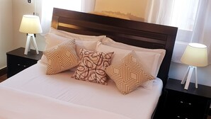 A neatly made bed with patterned pillows, two bedside tables with lamps, and sheer curtains. - Peaceful and secure Kyanja Oasis with Modern Amenities & Hot Shower (Kampala)