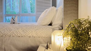 Quarto com cama, roupa de cama branca, mesa de cabeceira com abajur e janela com persianas.