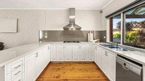 Cozinha moderna com armários brancos, coifa de aço inoxidável, lava-louças e janela com vista para vegetação.