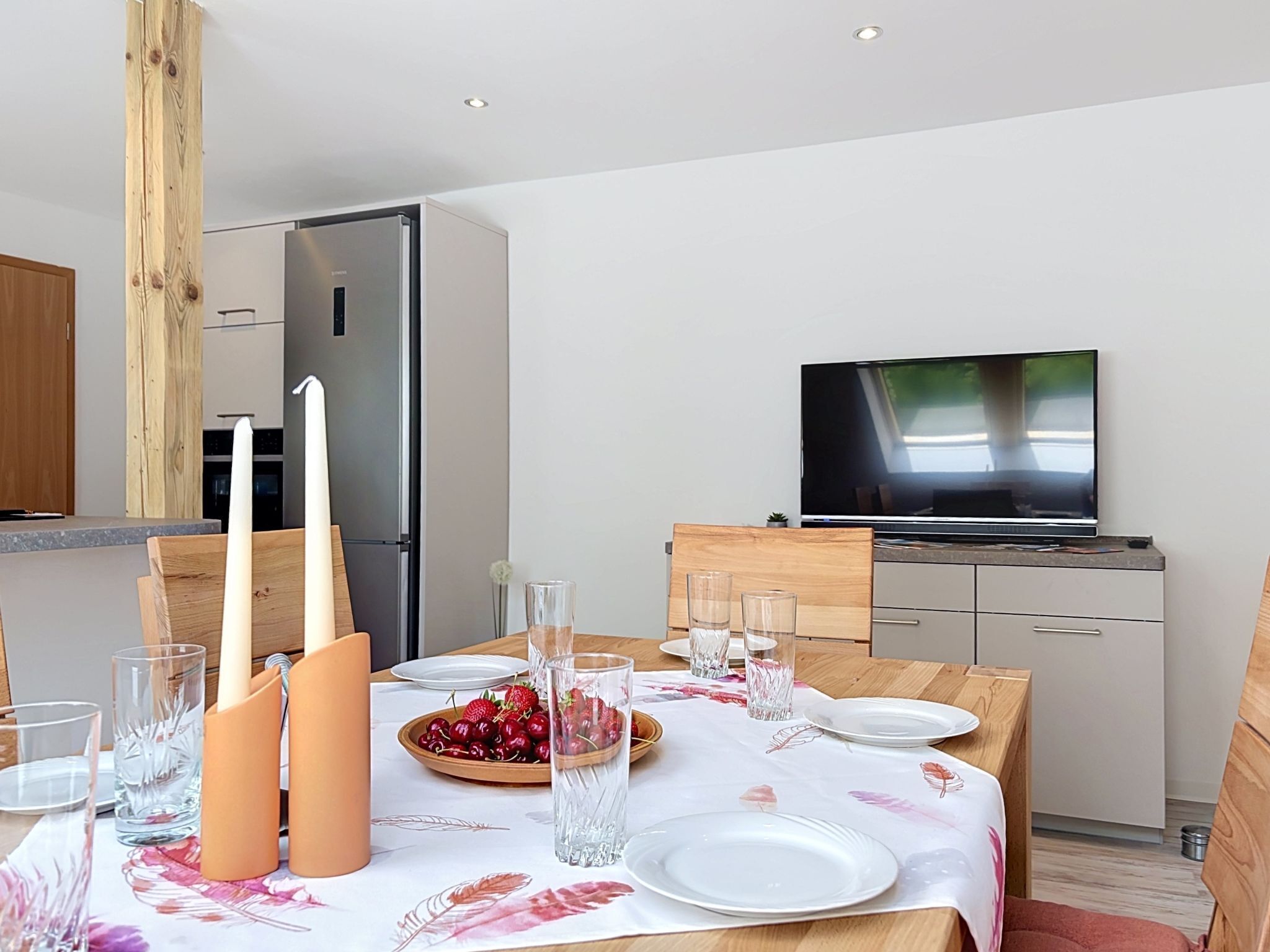 A modern dining area with a wooden table set for four, a TV mounted on the wall, and a kitchen in the background.