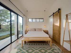 Une chambre à coucher avec un lit, deux tables de chevet, un banc en bois, une grande fenêtre et une vue sur l’extérieur.