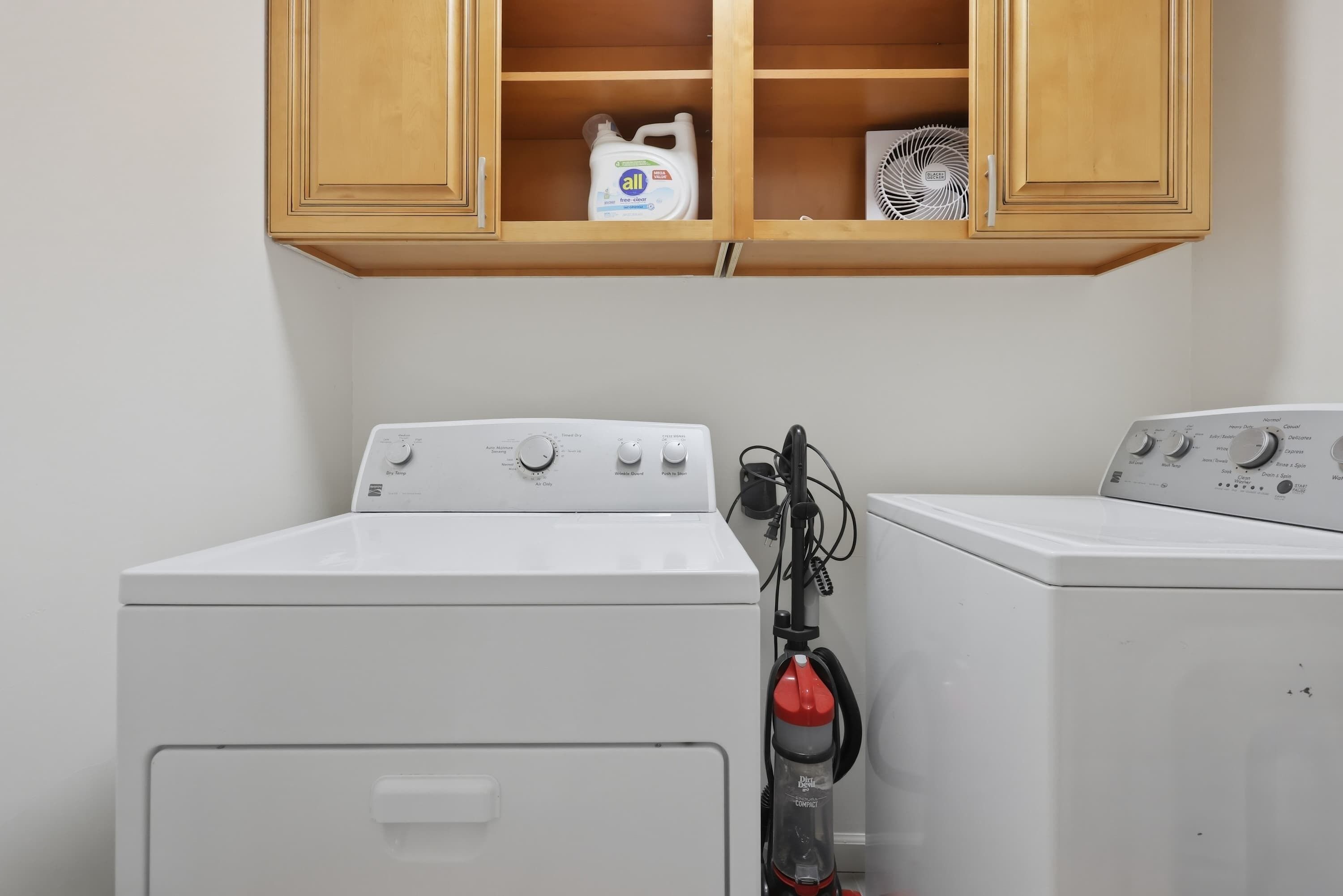 A laundry area with two white washing machines, a wooden shelf above containing a bottle of All detergent and a fan, and a red vacuum cleaner.