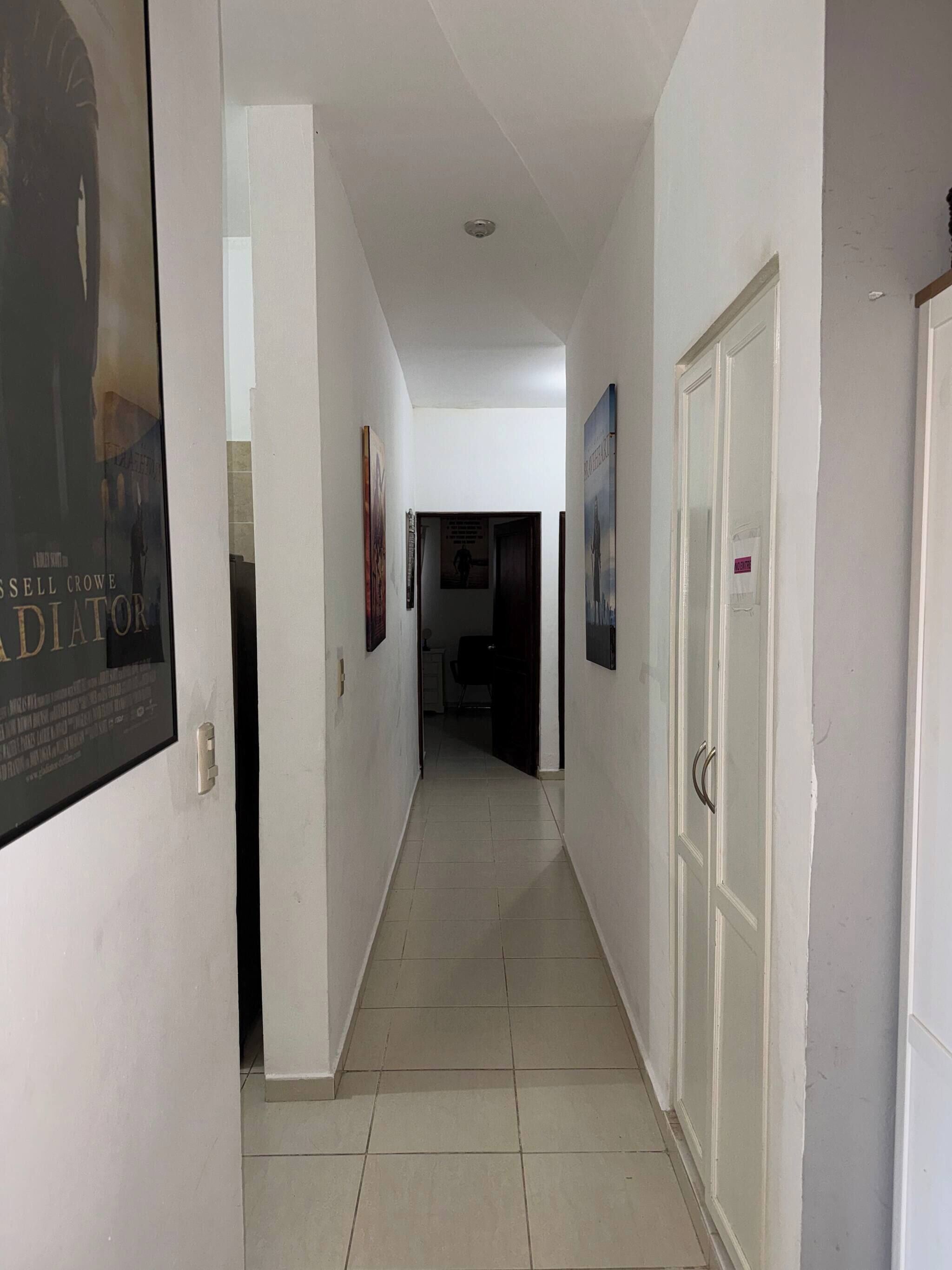 Un couloir aux murs blancs, doté d’un sol carrelé et d’une porte située sur le côté droit.