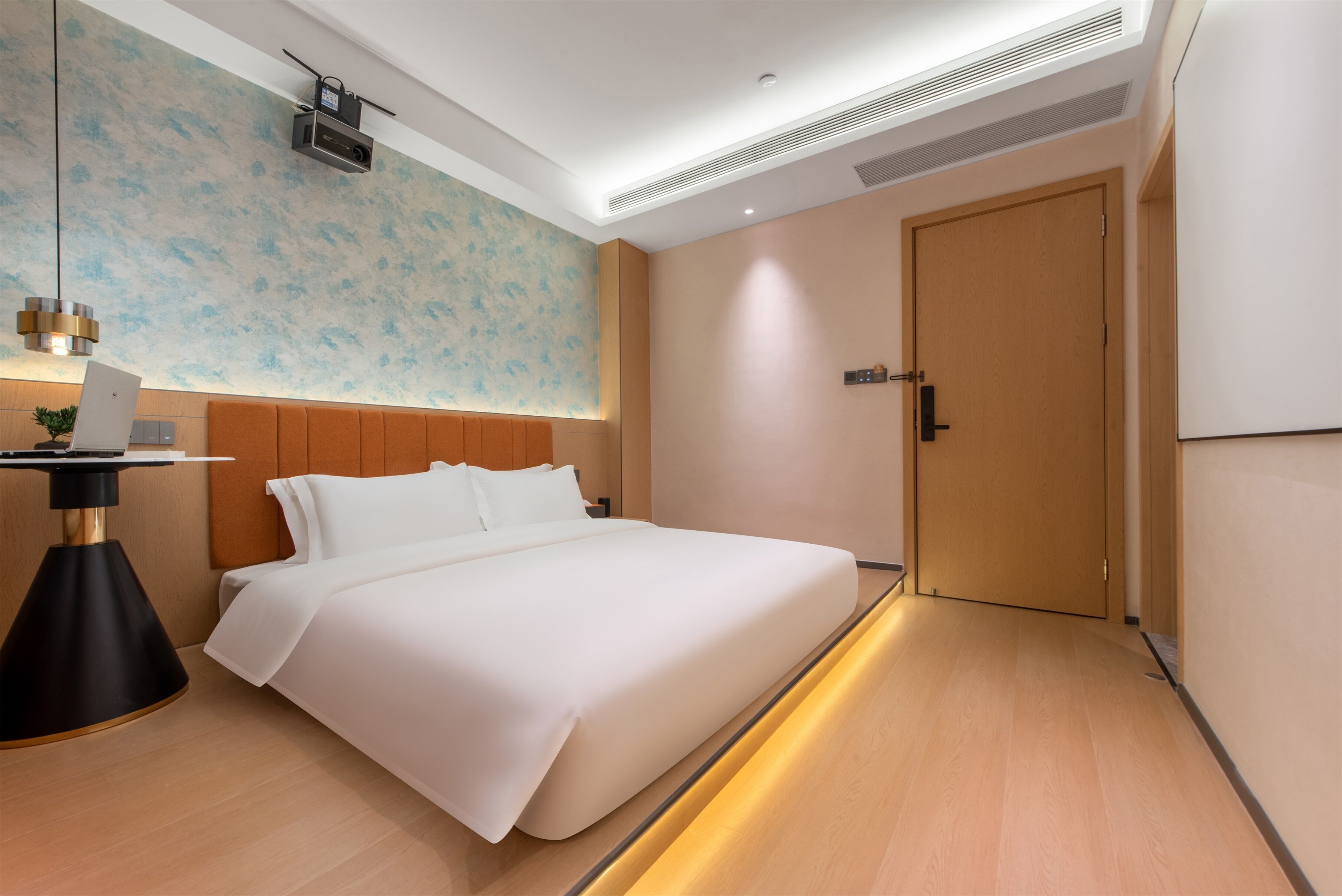 Una habitación de hotel moderna con una cama grande, un escritorio con lámpara, un proyector en la pared y una puerta de madera.