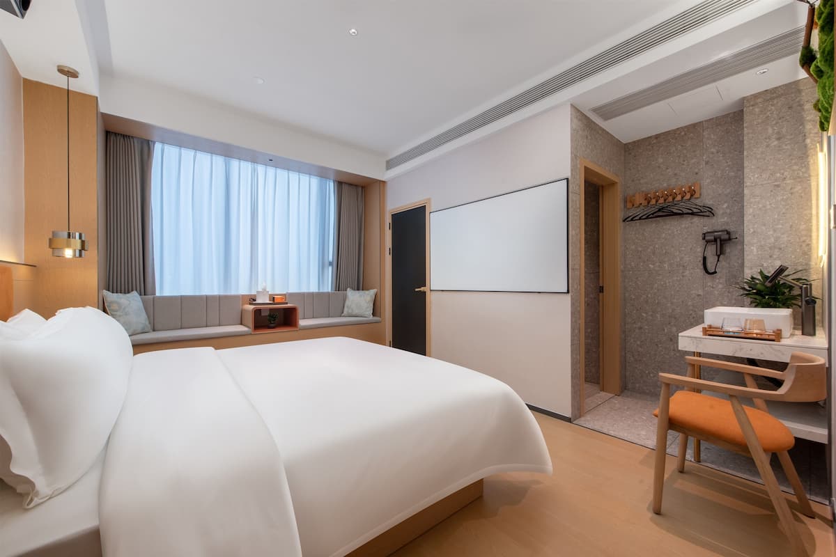 Ein modernes Hotelzimmer mit einem großen Bett, einem Schreibtisch mit Stuhl, einem großen Fenster mit Vorhängen, einem wandmontierten Fernseher und einer kleinen Pflanze.