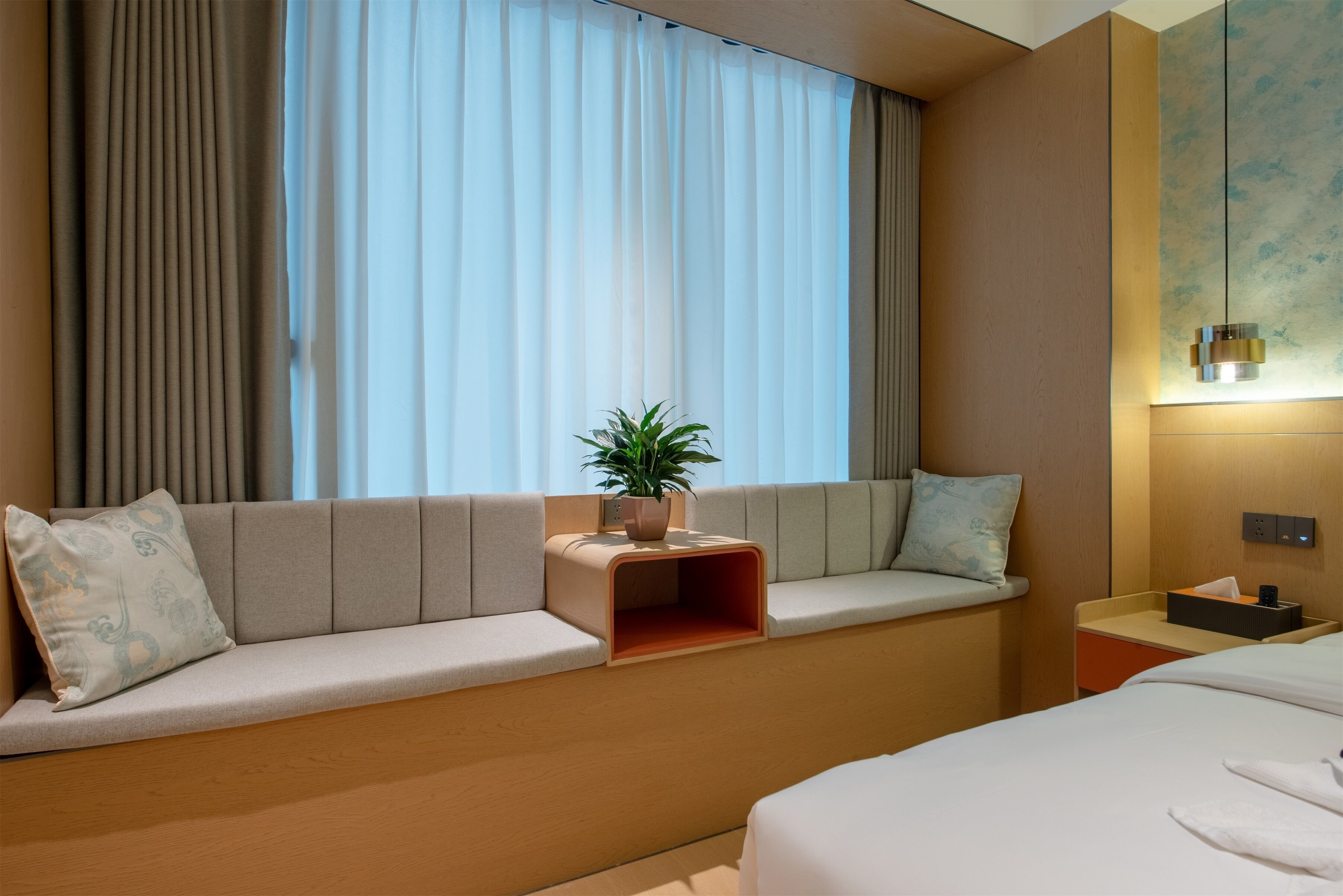 A hotel room with a sofa bed, a bedside table with a lamp, a small plant, and a window with curtains.