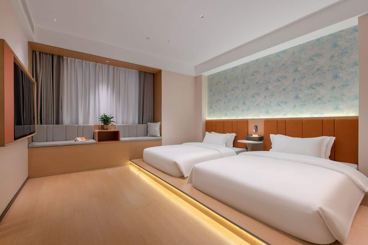Ein modernes Hotelzimmer mit zwei Betten, einer Sitzecke mit einer Pflanze, einem großen Fenster mit Vorhängen und einer Wand mit floralem Muster.