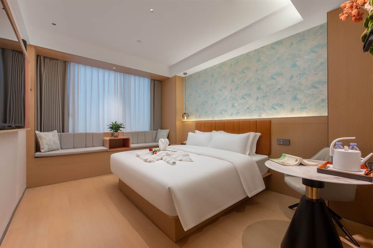 Ein modernes Hotelzimmer mit einem großen Bett, einem Nachttisch, einer kleinen Sitzecke, einem Fenster mit Vorhängen und einer Wand mit einem Blumenmuster.