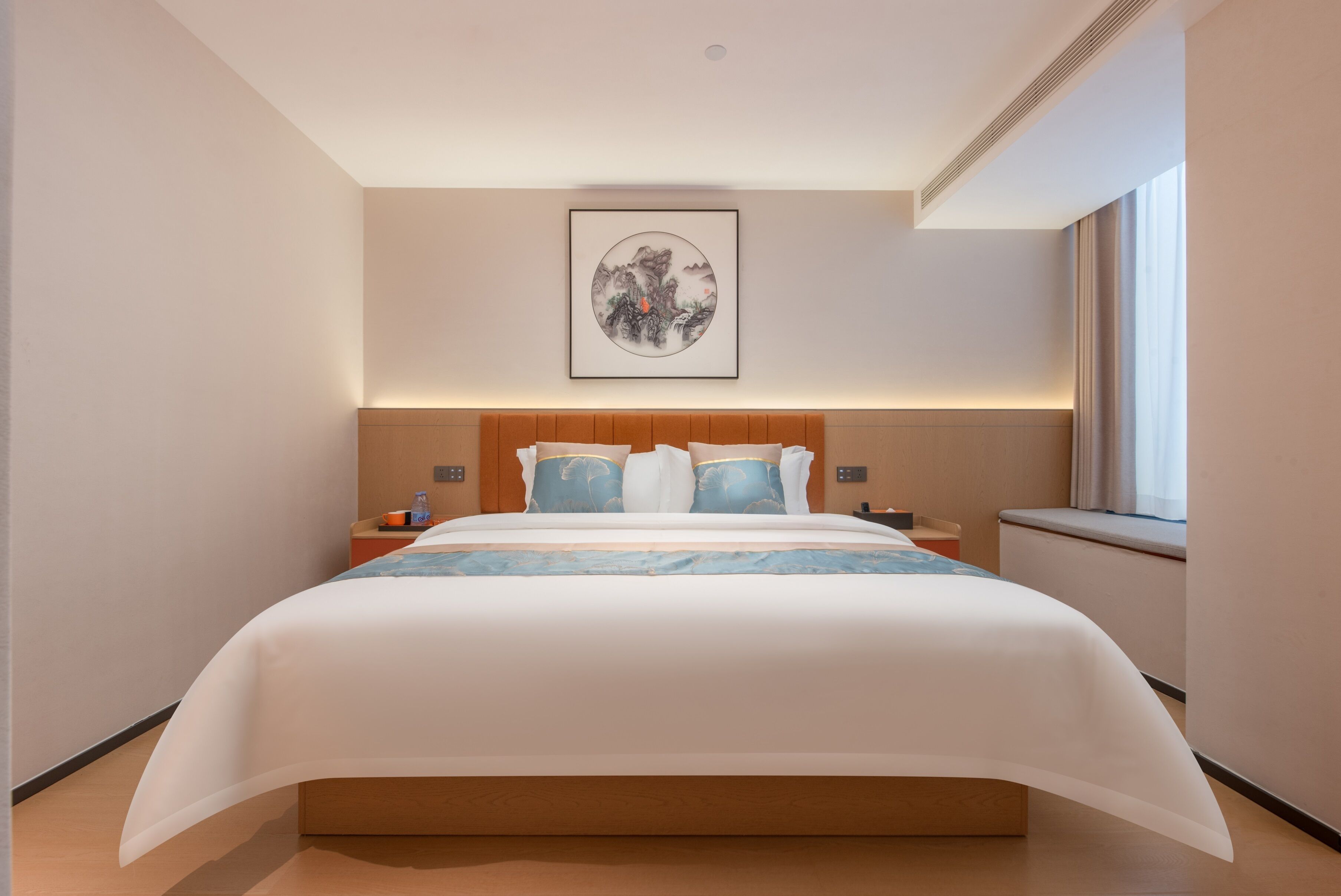 Une chambre d’hôtel moderne avec un grand lit, une tête de lit dotée d’un éclairage intégré, une œuvre d’art encadrée au mur et une fenêtre avec des rideaux.