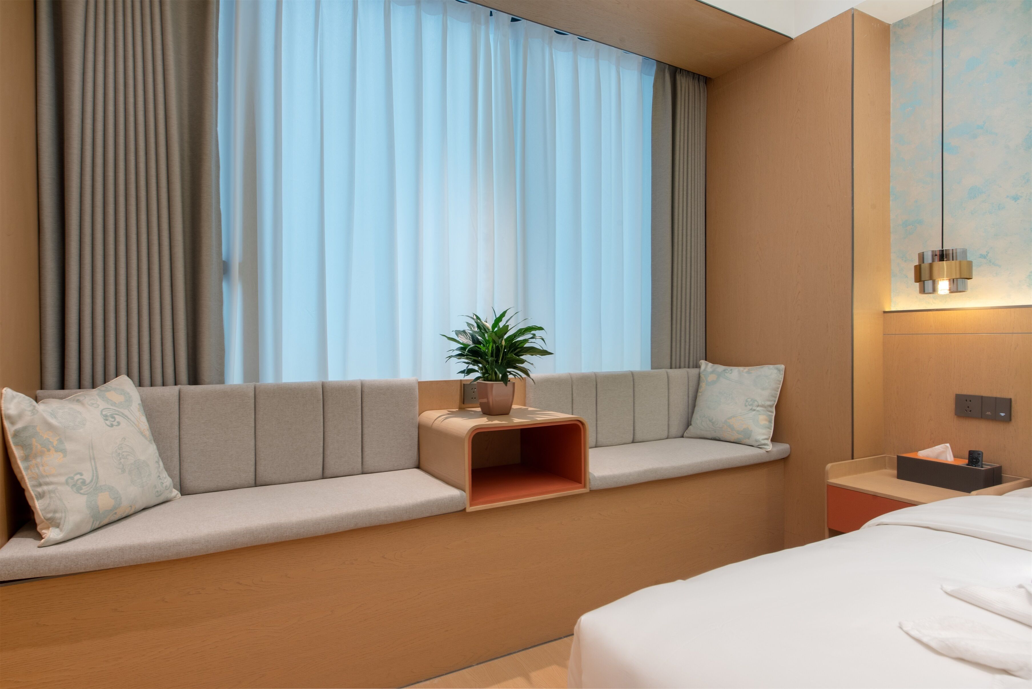 Ein modernes Hotelzimmer mit Schlafsofa, einem kleinen Tisch mit Pflanze, einem Nachttisch mit Lampe und einem großen Fenster mit Vorhängen.