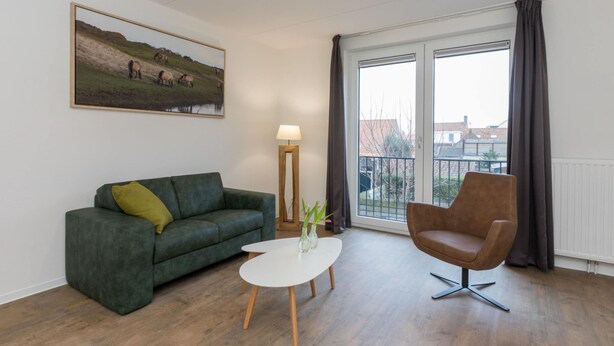 A living room with a green sofa, a white coffee table, a brown chair, a painting of horses, and a large window with curtains. - Apartment in Zoutelande Near Sunny Beach (Zoutelande)