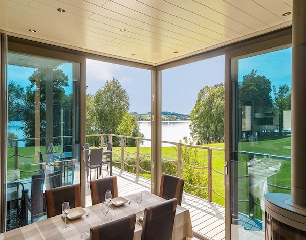 A dining area with a table set for four, overlooking a lake and a grassy area with trees.