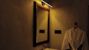 A bathroom with a sink, mirror, robe, and a can of toilet paper.