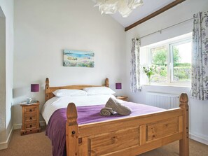 Un lit en bois avec une couvre-lit violette, une table de chevet avec une lampe, un tableau au mur et une fenêtre avec des rideaux.