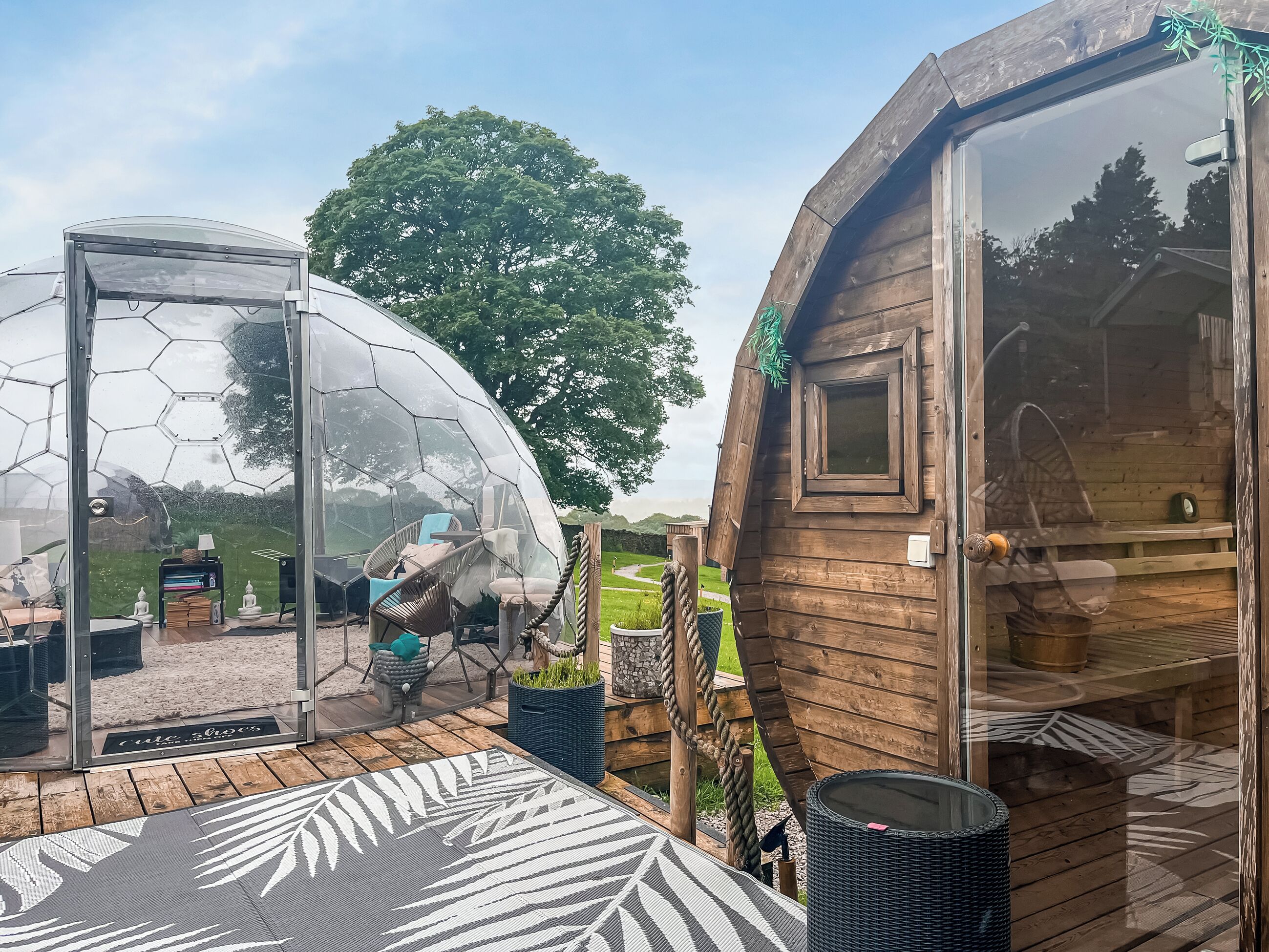 Uma estrutura de cúpula geodésica transparente com assentos no interior, adjacente a uma cabana de madeira com porta e janela.