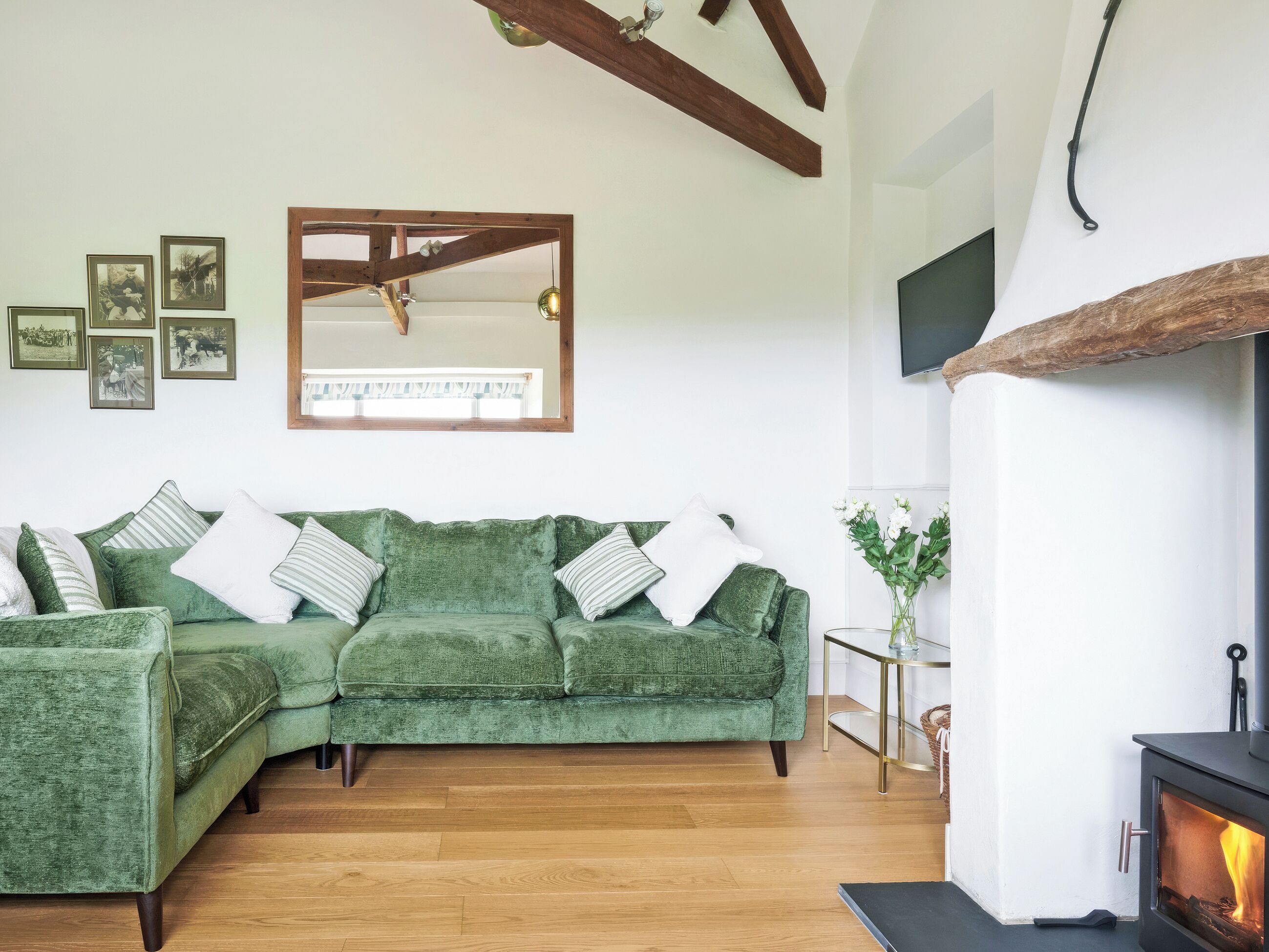 A green sofa with white and striped pillows, a wooden-framed mirror, a fireplace, and a small table with a vase of flowers.