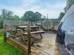 Una terraza de madera con mesas de picnic, una silla colgante y una parrilla.
