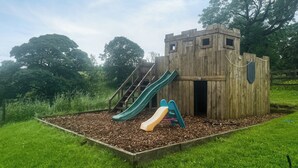 Eine hölzerne Spielanlage mit Rutschen und Treppen, umgeben von Gras und Bäumen.