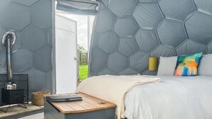 Ein Schlafzimmer mit einem Bett, einem Heizgerät, einer Holzkiste und Blick durch eine offene Tür.