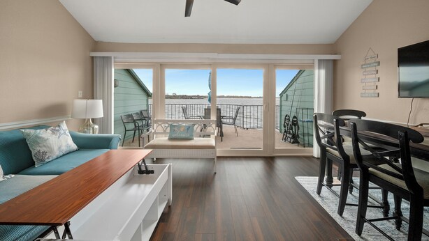 A living room with a sofa, coffee table, and dining table, a ceiling fan, and a view of the ocean through large windows. - Fishing on the Overwater Balcony! 2BR/2BA Condo with Beautiful Lake Conroe View (Willis)