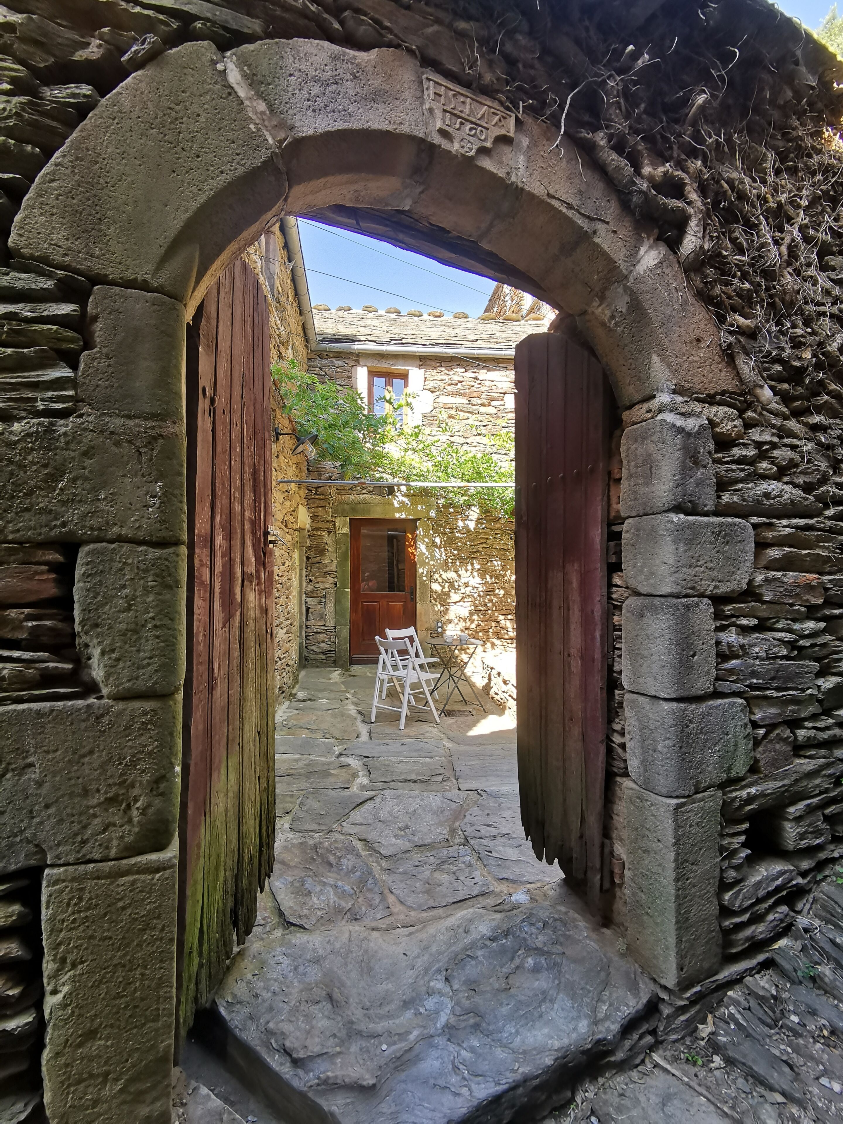 A stone archway leading to a rustic stone building with a wooden door and a small table with chairs outside.