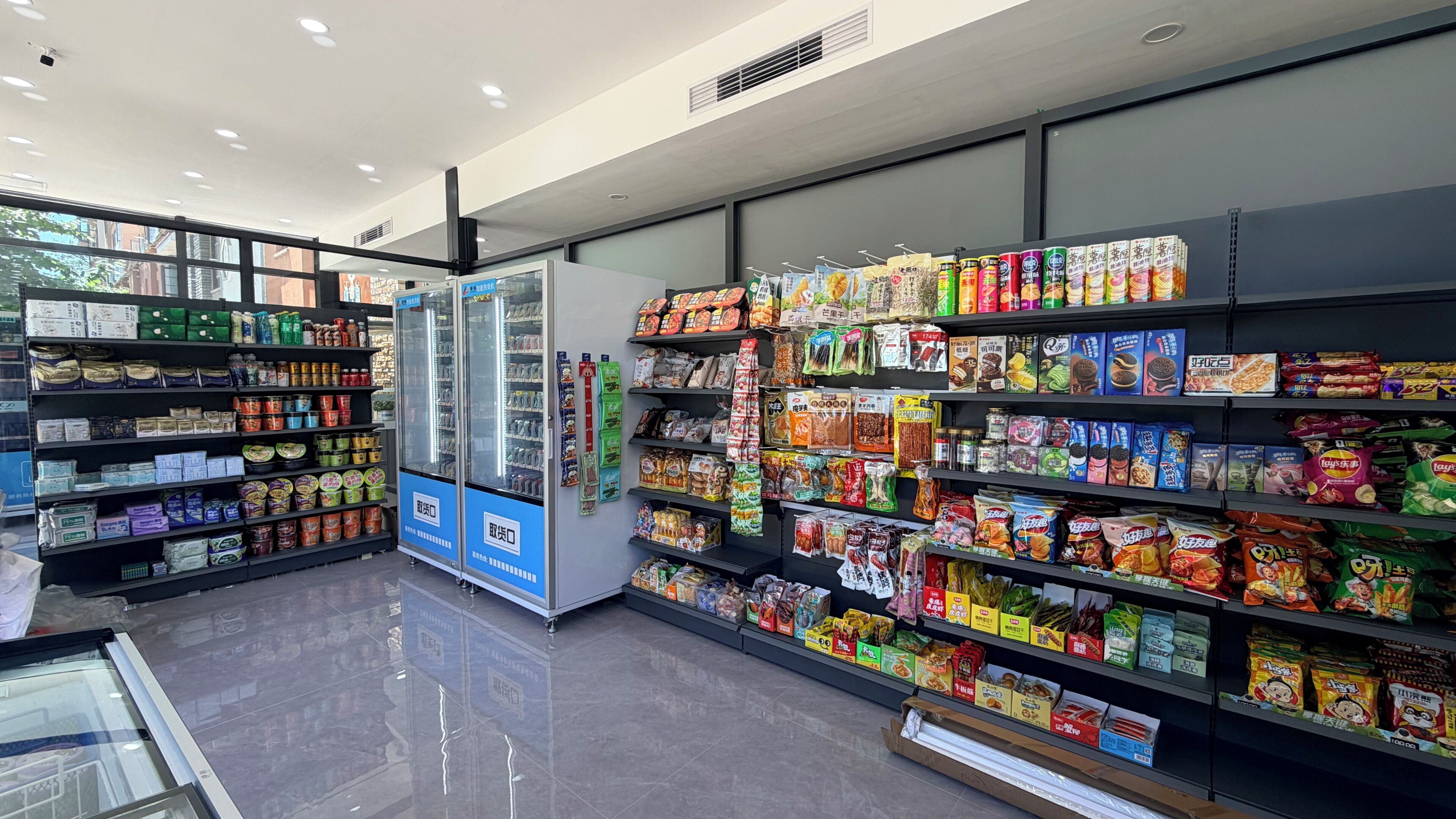 Interior de una tienda de comestibles con estantes llenos de diversos alimentos, un refrigerador y una máquina expendedora.