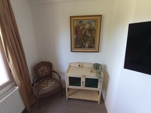 A room with a radiator, a chair with floral upholstery, a small white cabinet with a green plant, and a framed painting on the wall. - Müllerstube im Müllerhaus (Lübeck)