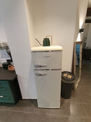 Ein kompakter, weißer Kühlschrank mit grünem Oberteil in einer Küche mit Fliesenboden und Spiegel an der Wand.