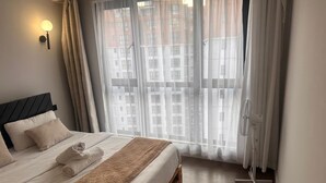 Ett hotellrum med en säng, en väggmonterad lampa, ett fönster med gardiner och en golvfläkt.