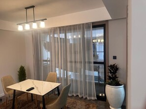 A modern room with a dining table, chairs, a large potted plant, and a view through sheer curtains. - 
Heated POOL 1Bedroom Fully Equipped GYM Balcony view of Nairobi National Park (Machakos)
