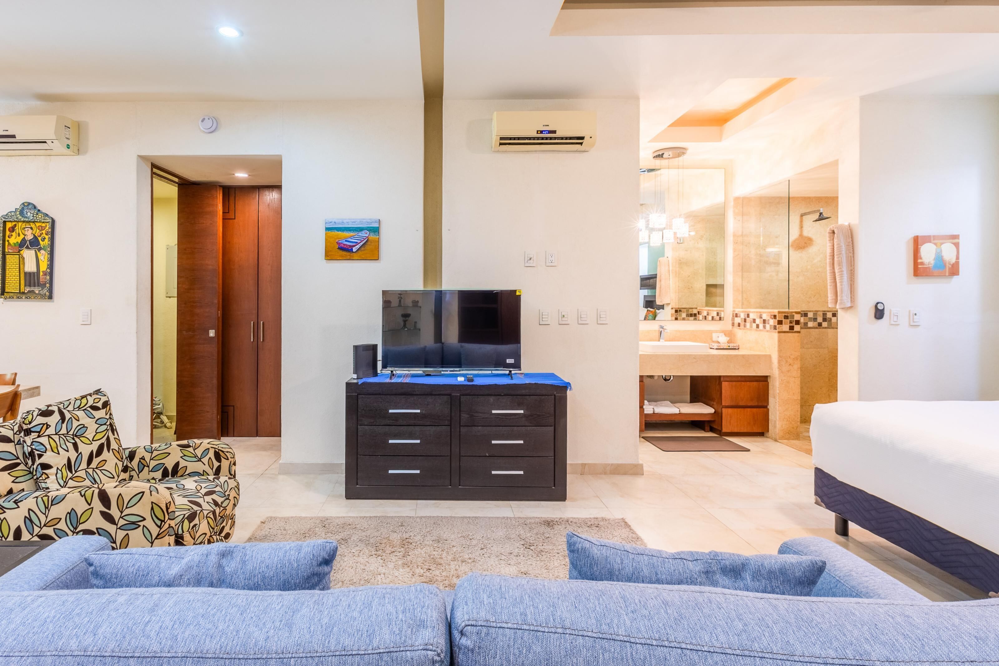 A modern hotel room with a sofa, TV, minibar, and a bathroom with a shower and sink.