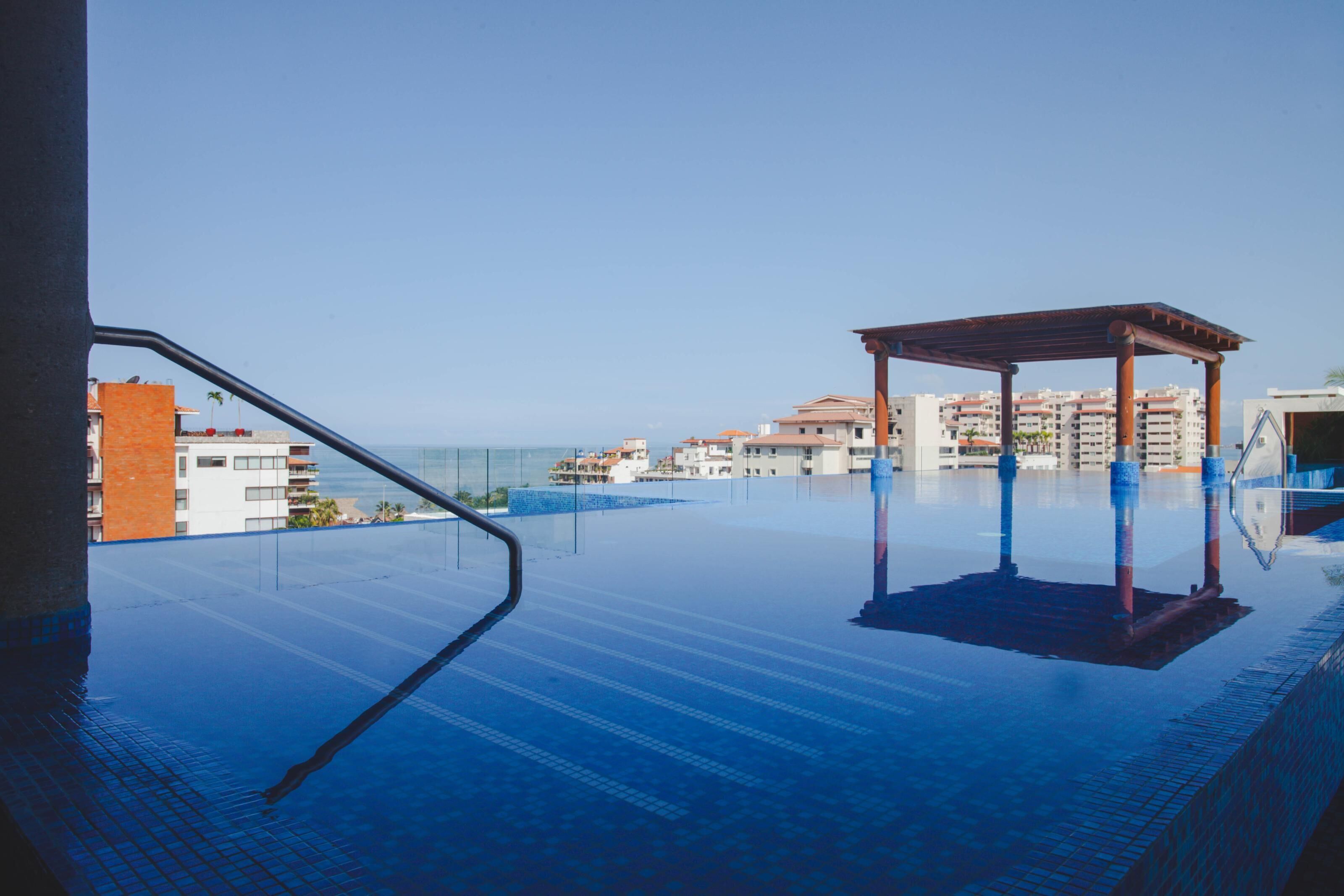 An infinity pool with a wooden pergola, clear sky, and buildings in the background.
