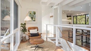 A spacious room with a high ceiling, a large window, a brown leather chair, a white lamp, and a plant. - Maisie's by Holiday Management Co. (Bega Valley)