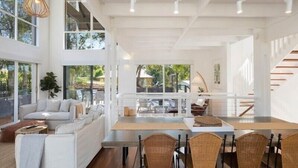 A spacious living area with a sofa, dining table, and chairs, large windows, and a staircase. - Maisie's by Holiday Management Co. (Bega Valley)