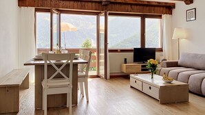 Quarto com teto de madeira, mesa de jantar com cadeiras, sofá, televisão e janelas amplas com vista para as montanhas.