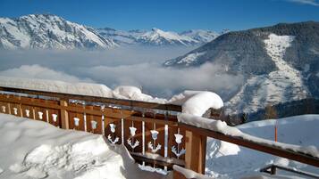 Une clôture en bois, recouverte de neige, surplombe un paysage montagneux aux sommets enneigés et à la vallée qui s’étend en contrebas.