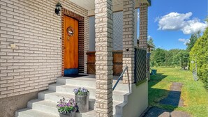 Uma casa de tijolos com porta de madeira, degraus que levam à entrada e um jardim com gramado e árvores.