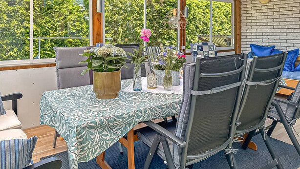Uma sala de jantar com uma mesa posta para quatro pessoas, rodeada por cadeiras, e vista para o verde através de amplas janelas.