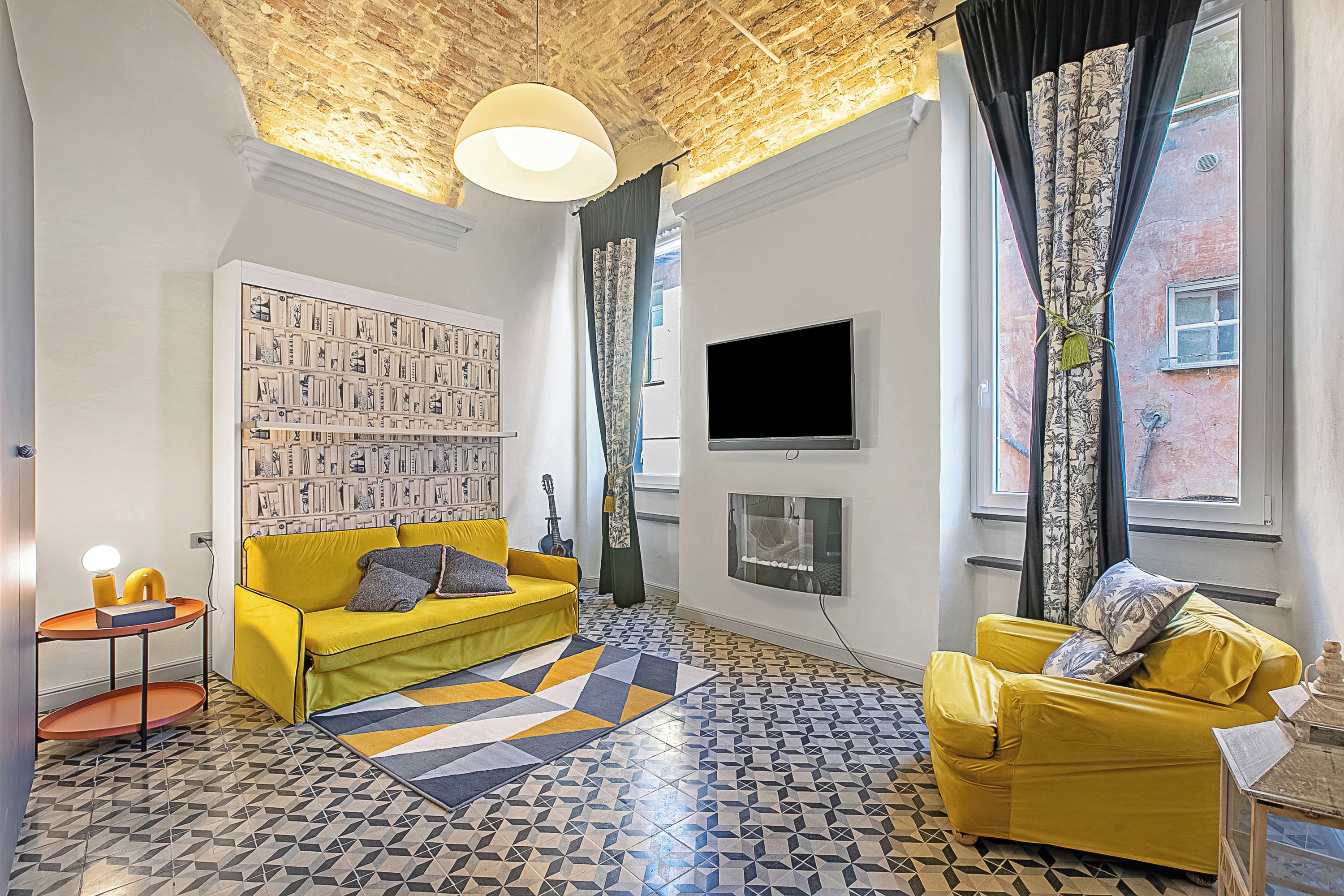 A living room with a yellow sofa, a patterned rug, a wall-mounted TV, and a bookshelf.