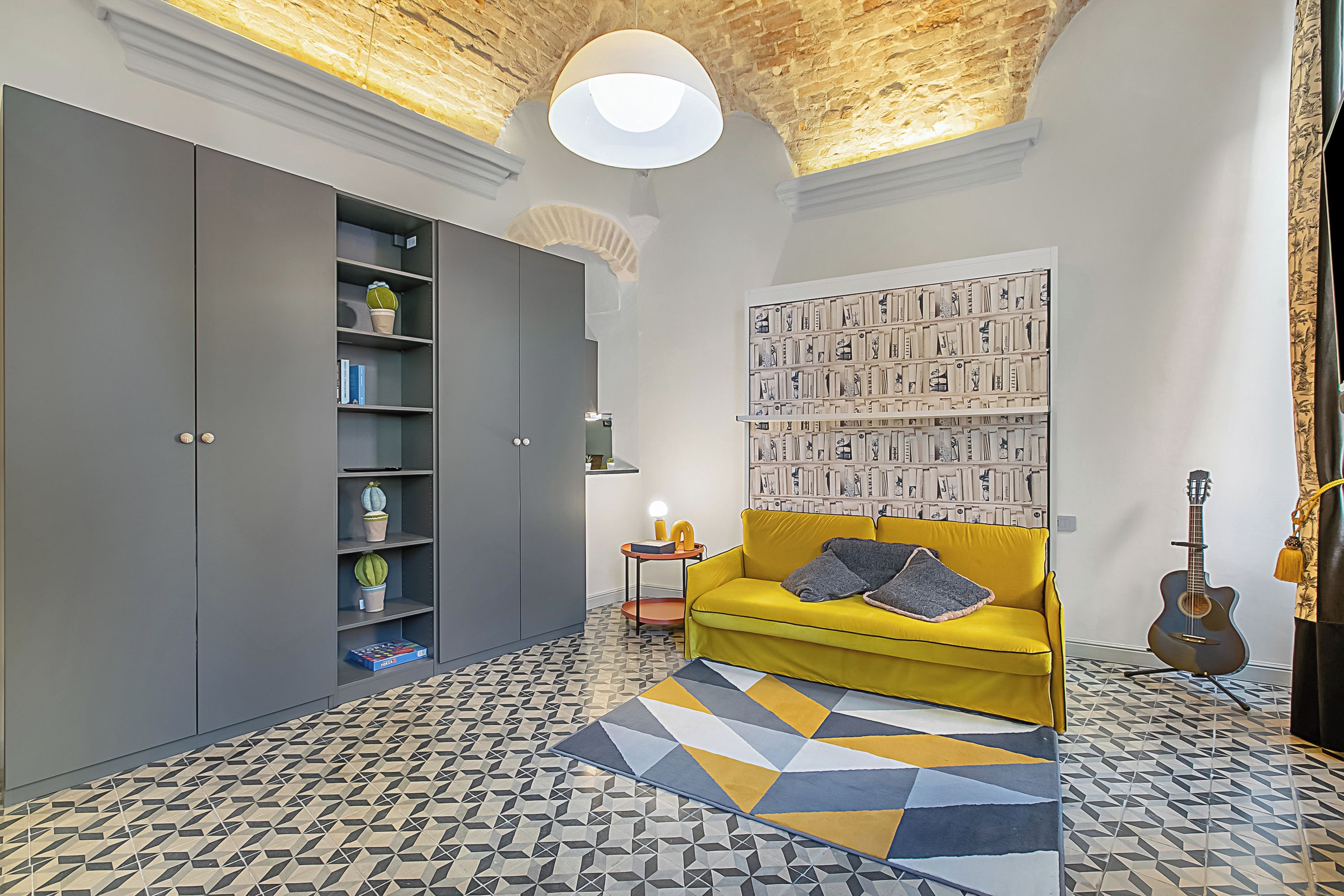 A room with a yellow sofa, a bookshelf, a guitar, and patterned flooring.