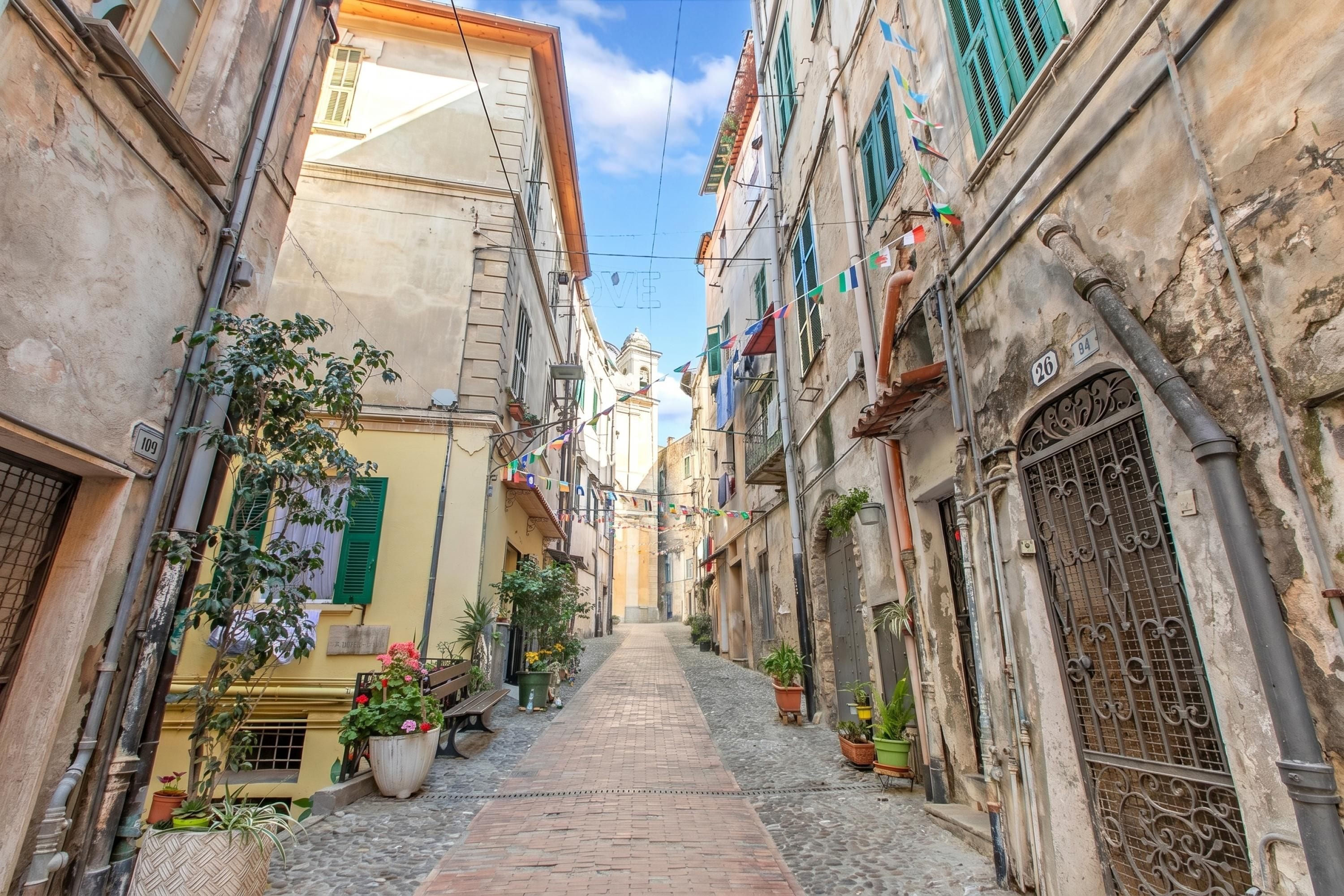 Une ruelle étroite pavée, bordée de bâtiments historiques dotés de balcons, de plantes en pot et surplombée d’un ciel dégagé.
