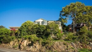 Une maison dotée d’une cheminée imposante, entourée d’une végétation luxuriante et située au pied d’une colline rocheuse.