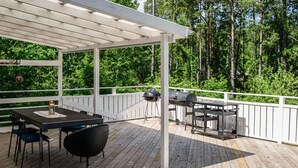 En takförsedd uteplats med ett bord och stolar, en grill och utsikt över en skog.