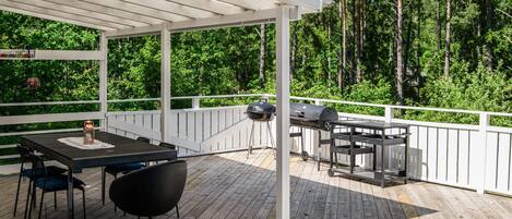 A covered outdoor deck with a table and chairs, a grill, and a view of a forest.