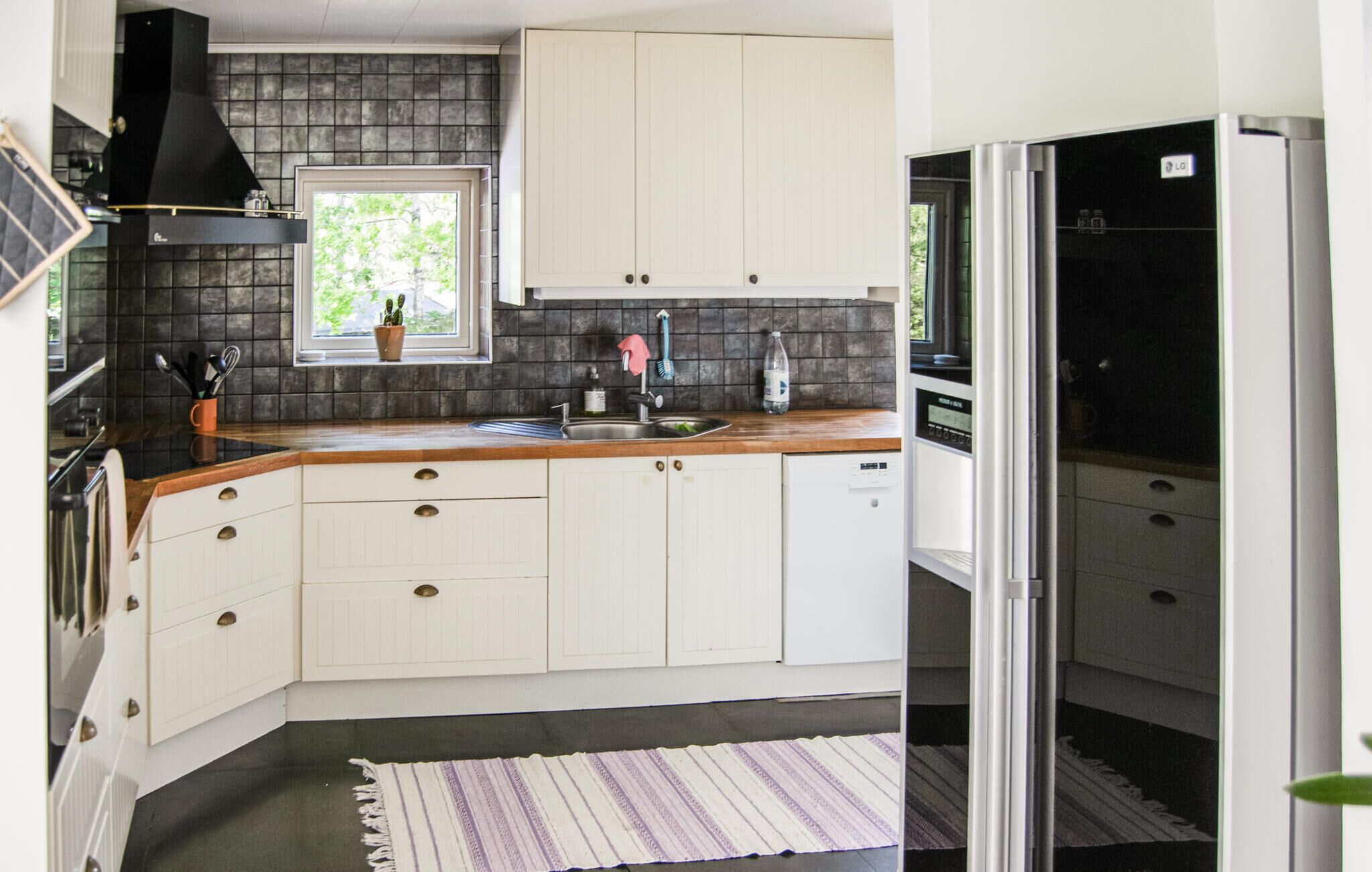 A compact kitchen with white cabinets, a black range hood, a window with a plant, and a striped rug.