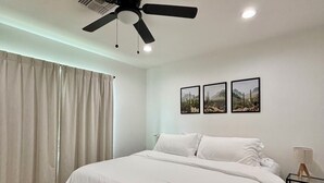 Une chambre à coucher avec un lit, un ventilateur de plafond, une fenêtre avec des rideaux et trois tableaux encadrés accrochés au mur.