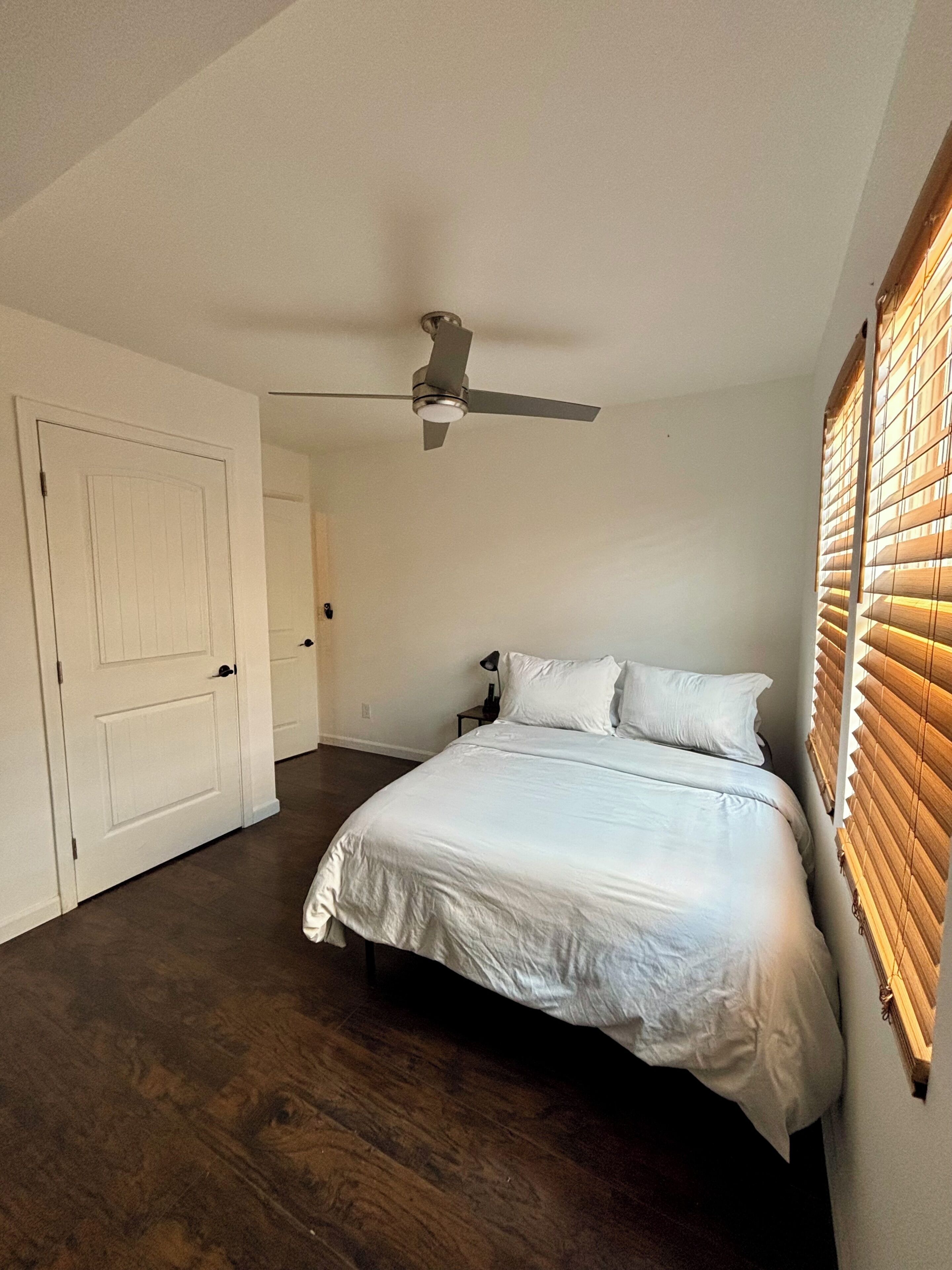 A bedroom with a bed, a ceiling fan, a door, and a window with blinds.