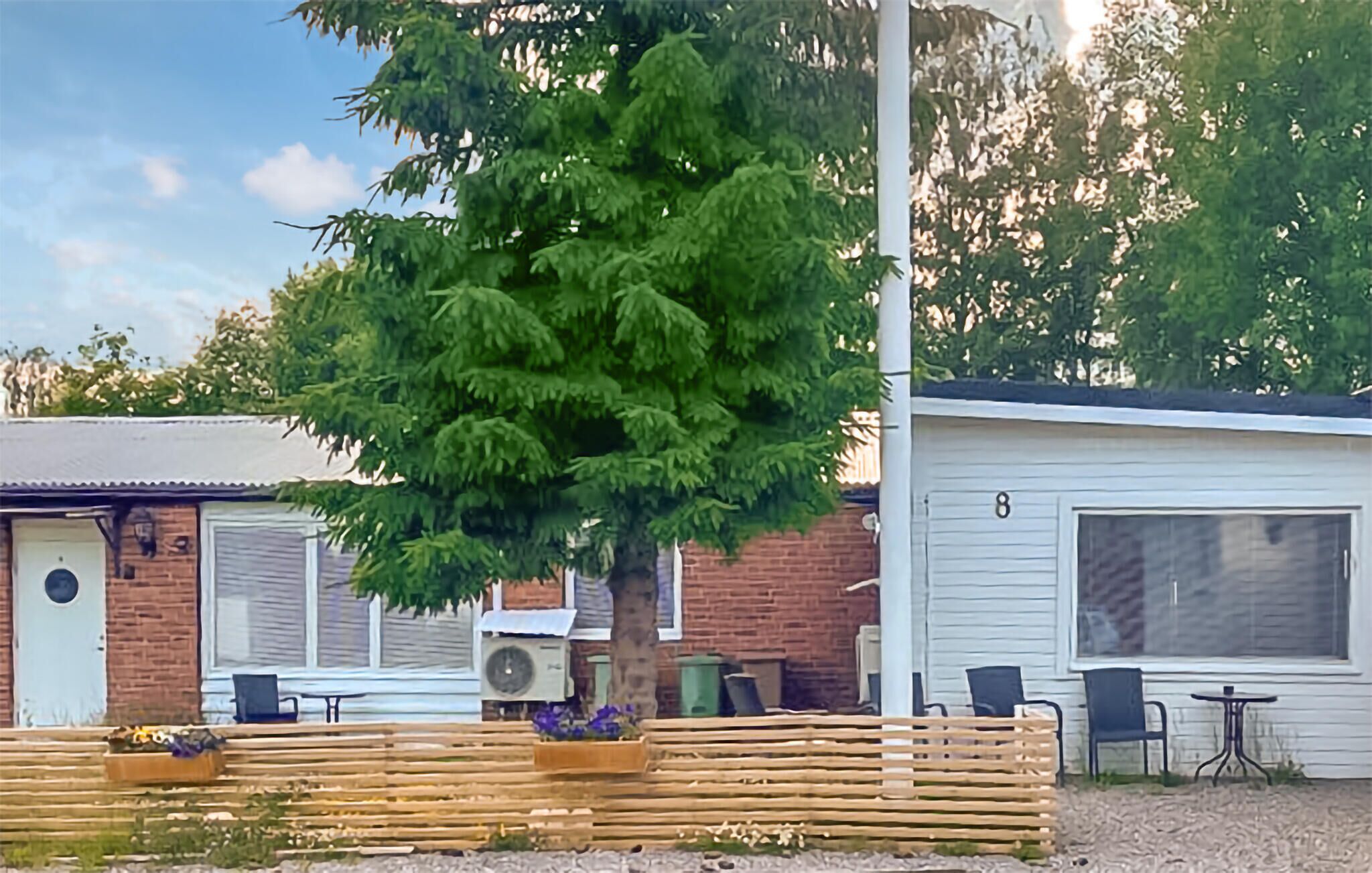 Ein eingeschossiges Gebäude mit Ziegelaußenwand, ein großer Baum davor und eine kleine Terrasse mit Gartenmöbeln.
