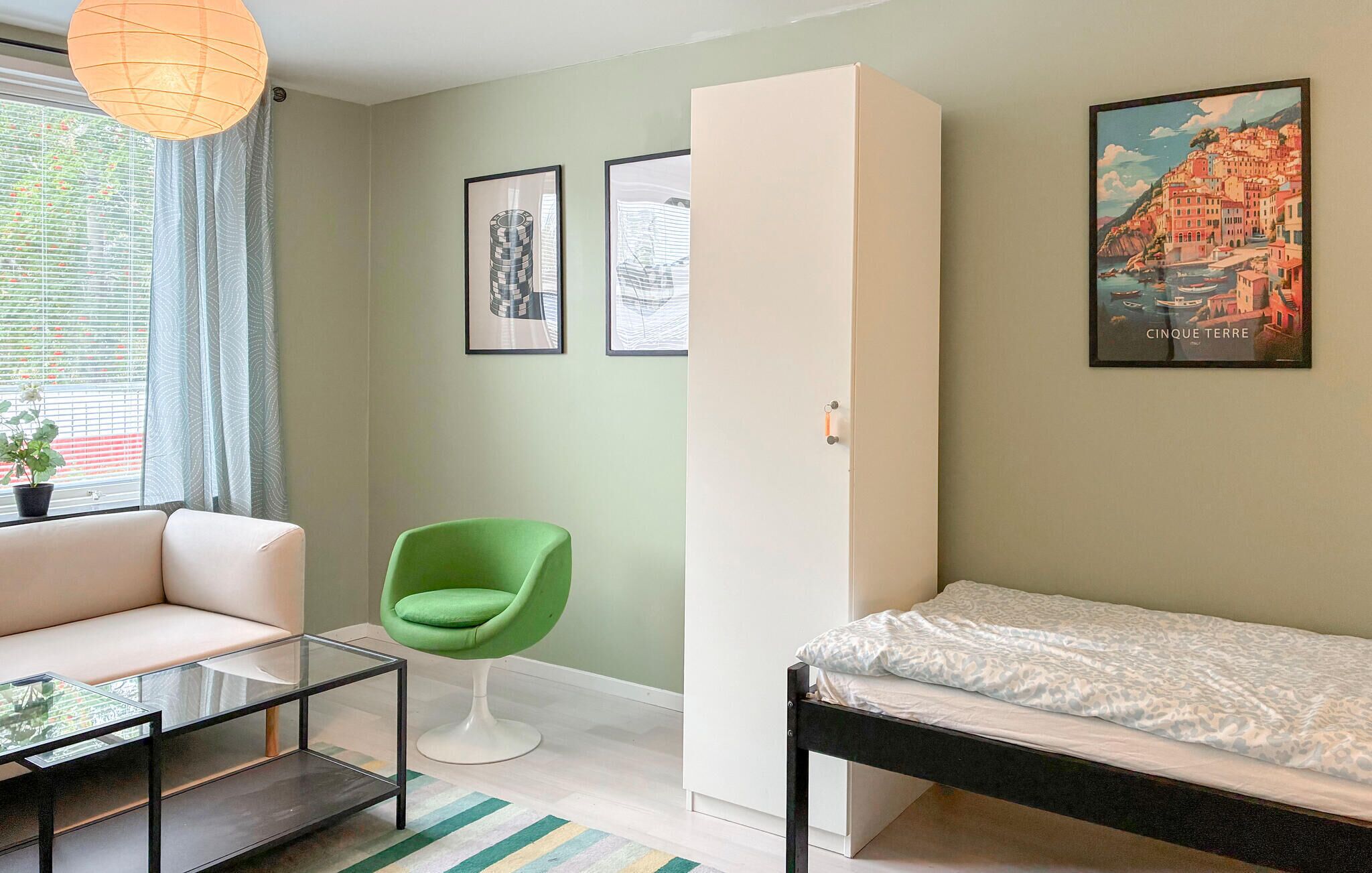 Een kamer met een bank, een groene fauteuil, een bed, een glazen salontafel en twee ingelijste foto’s aan de muur.