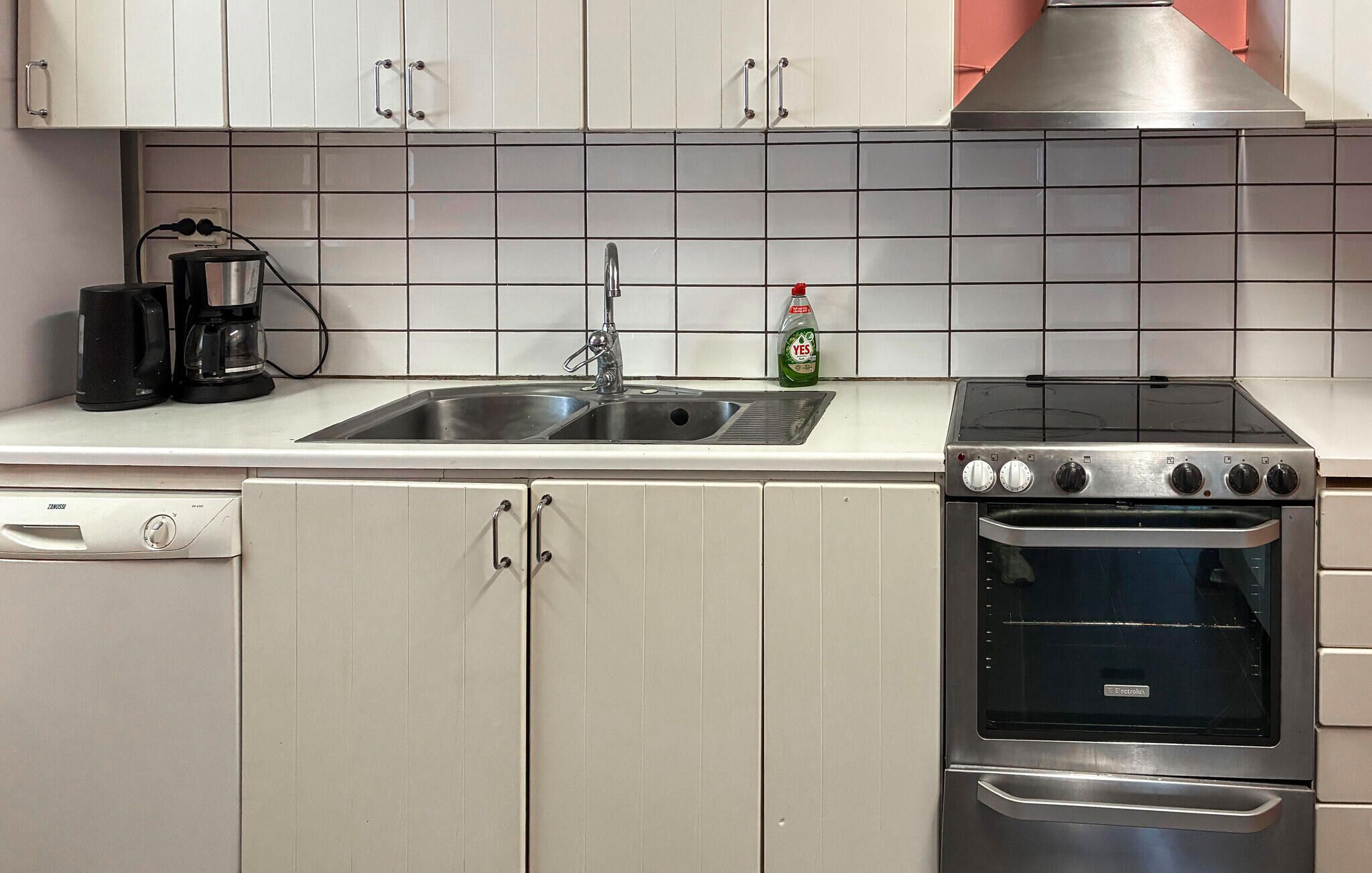 Een keuken met een dubbele spoelbak, een koffiezetapparaat, een fles afwasmiddel en een roestvrijstalen fornuis.